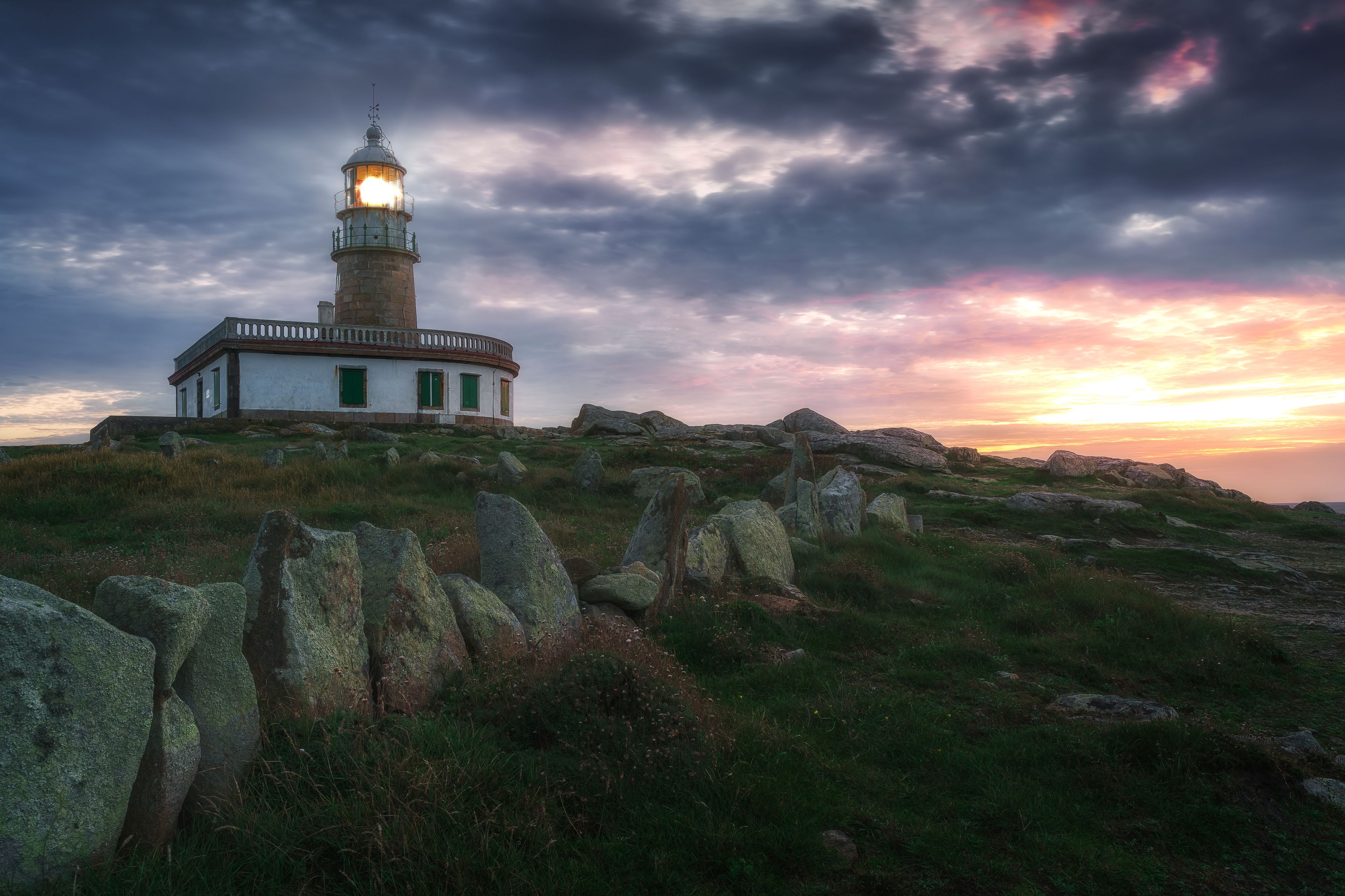A lighthouse on a grassy hill with large rocks in the foreground and a sunset in the background.