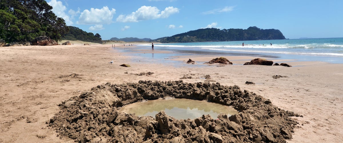 Una spiaggia sabbiosa con una formazione rocciosa circolare in primo piano e una collina boscosa sullo sfondo.