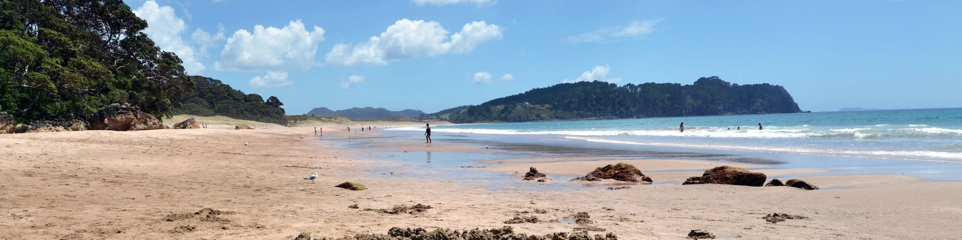 A sandy beach with a circular rock formation in the foreground and a forested hill in the background.