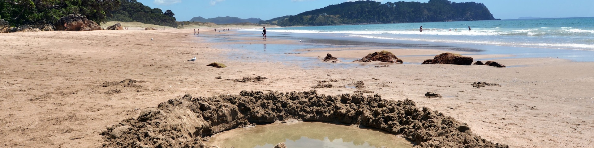 Ein sandiger Strand mit einer runden Felsformation im Vordergrund und einer bewaldeten Hügelkette im Hintergrund.