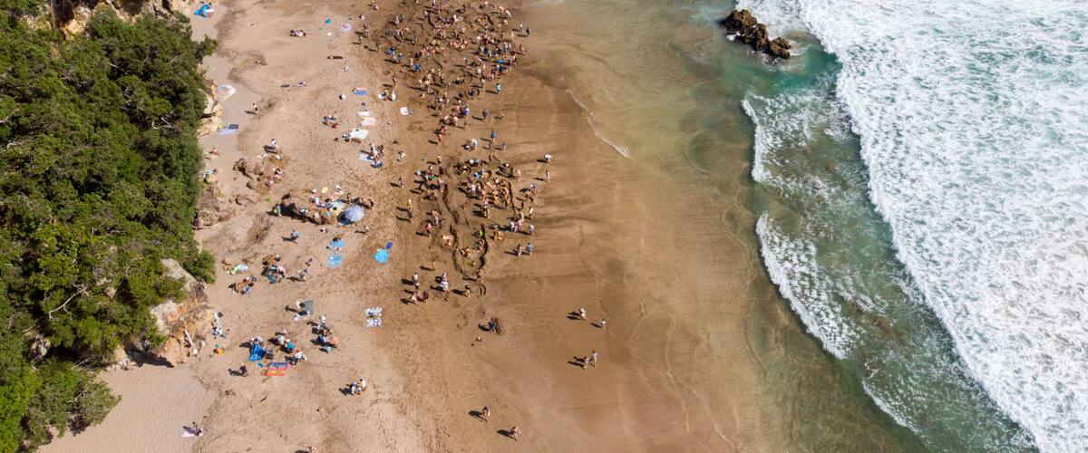 An aerial view of a beach with people, tents, and a rocky outcrop near the water.