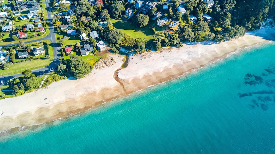An aerial view of a coastal area with a sandy beach, clear turquoise waters, and a residential neighborhood with houses and greenery.