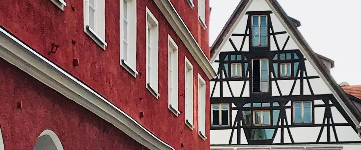 Un edifici vermell amb emmarcadures de finestres blanques i un edifici de fusta amb fustes fosques i clares al fons.
