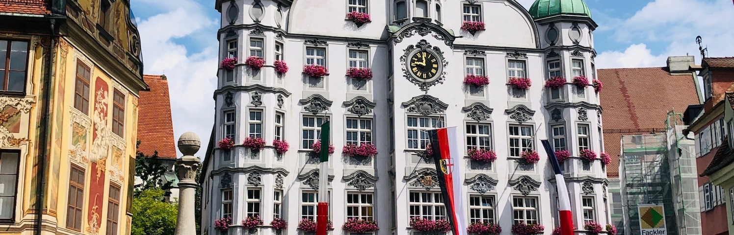 A historic building with green domes and red flowers, a clock, and flags.