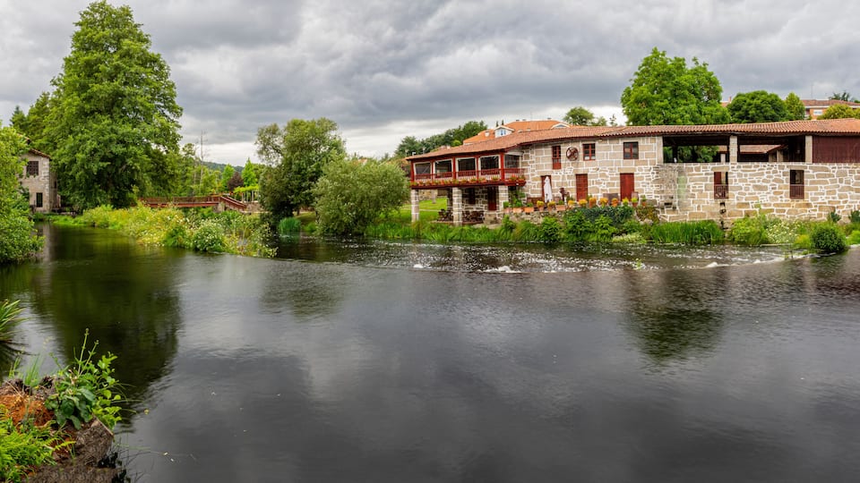 A riverside building with a stone facade and red accents, surrounded by lush greenery and trees.