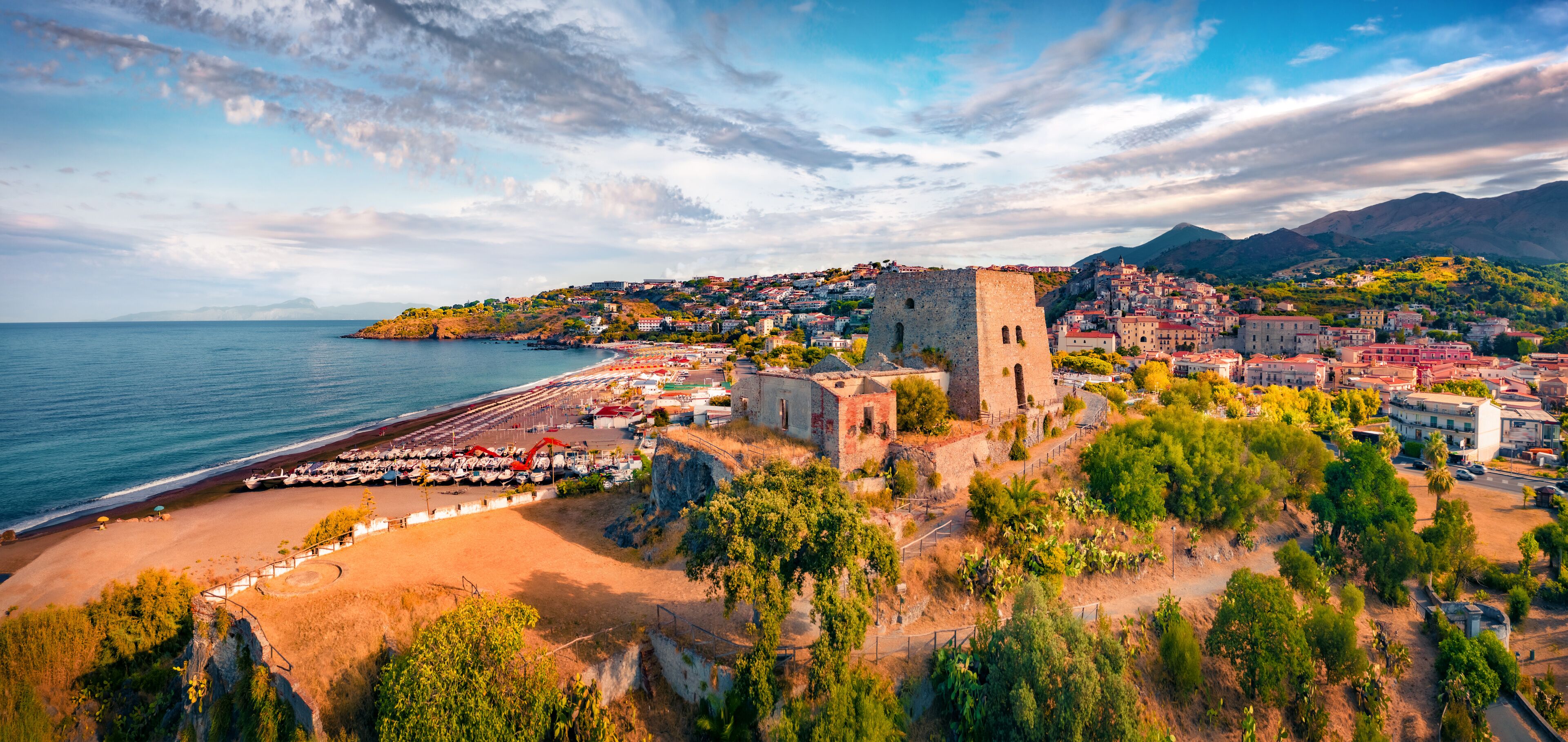 Un pueblo costero con una torre histórica, una playa con coches aparcados y montañas al fondo.