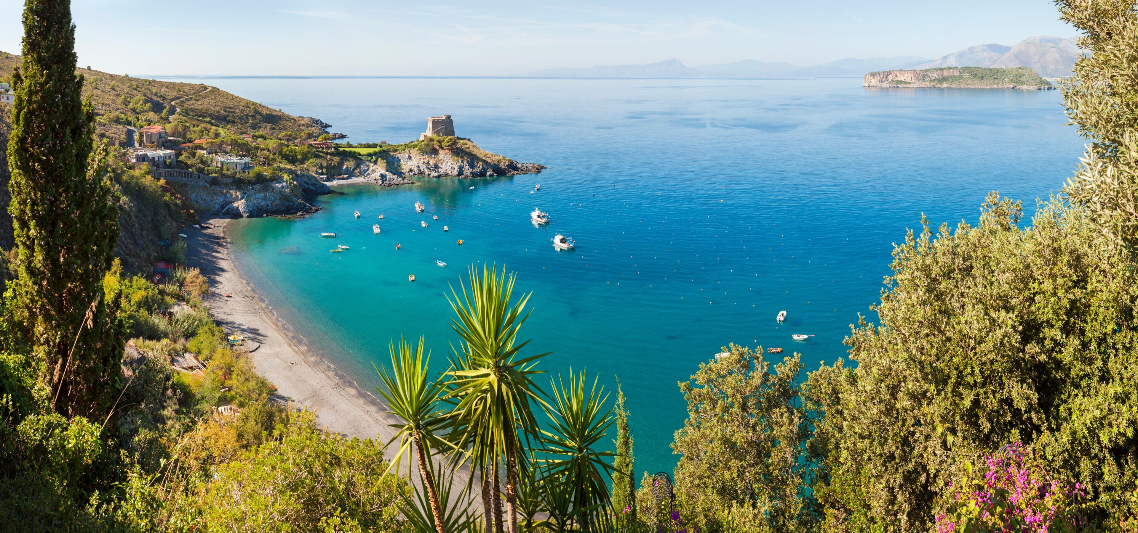 A coastal landscape with a clear blue sea, boats, a beach, and a historic tower on a cliff.