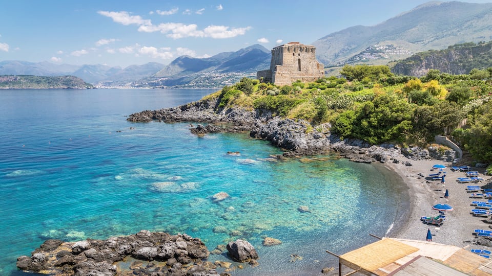 A coastal landscape with clear turquoise waters, a rocky shoreline, a historic tower, and a beach with umbrellas.