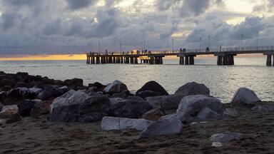 A pier extending into the water with people fishing, surrounded by large rocks and a cloudy sky.