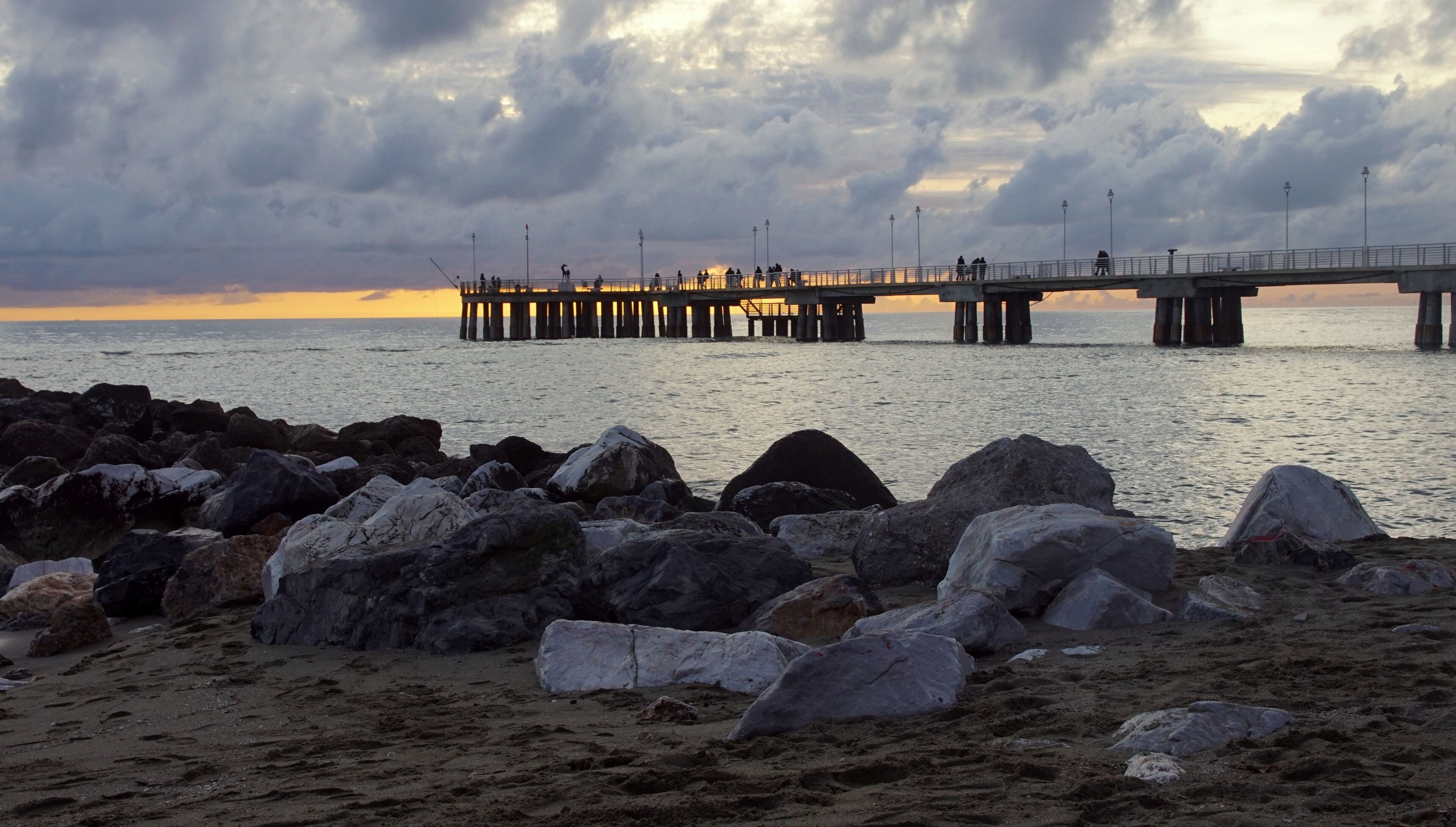 A pier extending into the water with people fishing, surrounded by large rocks and a cloudy sky.