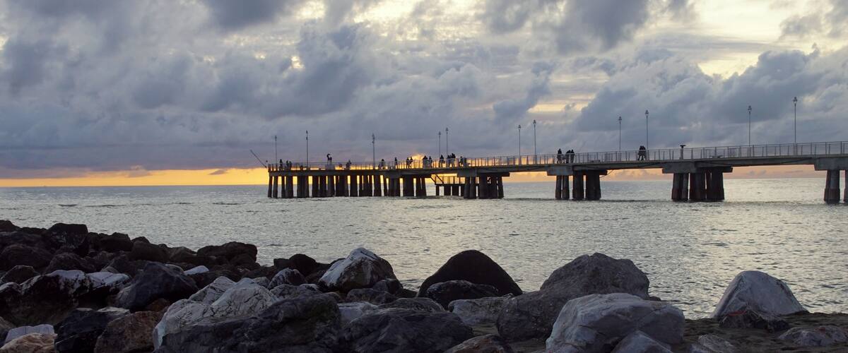 A pier extending into the water with people fishing, surrounded by large rocks and a cloudy sky.