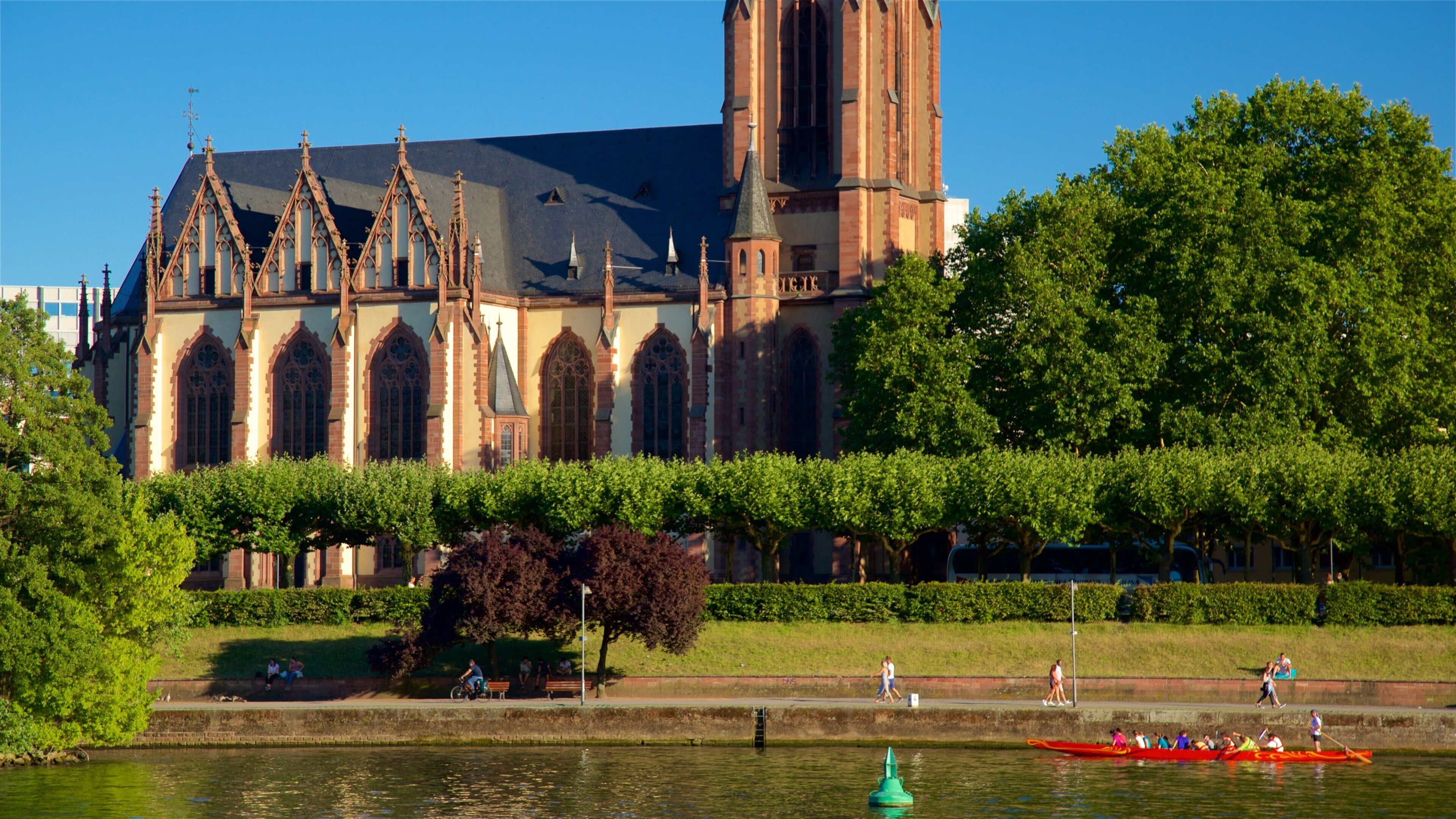 A historic church with Gothic architecture, a river with a rowboat, and a park with people.