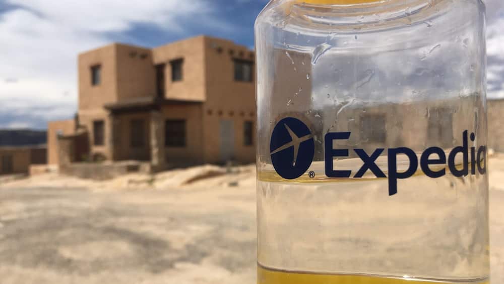 A clear glass bottle with a yellow liquid, labeled 'Expedia', in front of a desert landscape with buildings.