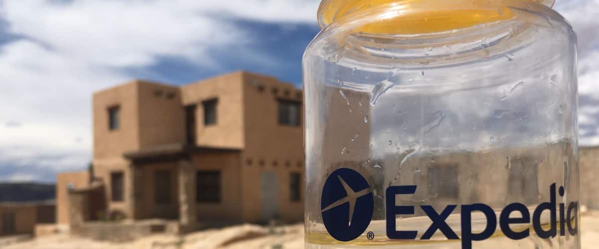 A clear glass bottle with a yellow liquid, labeled 'Expedia', in front of a desert landscape with buildings.