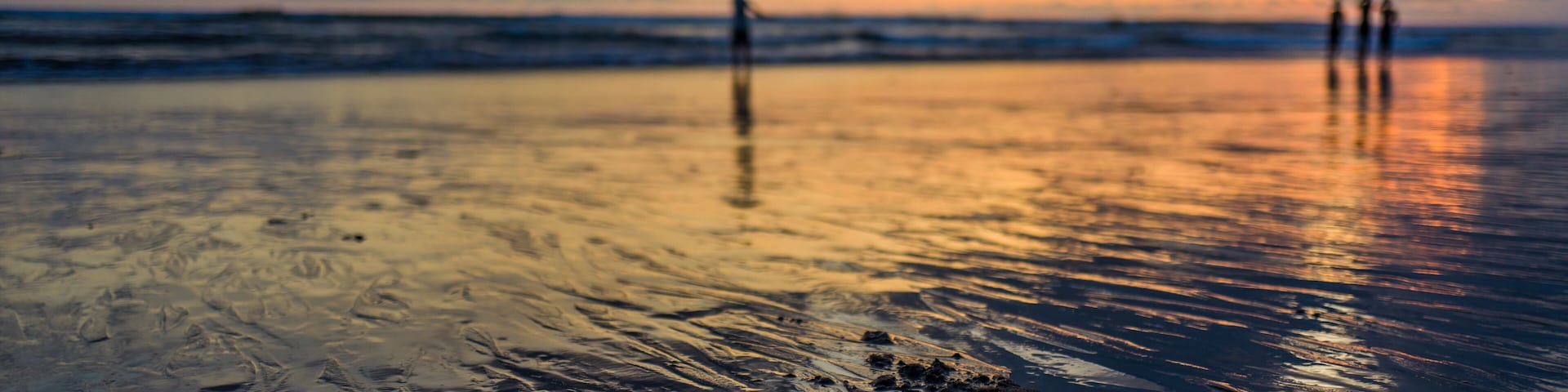 Una playa al atardecer con olas, cielo nublado y algunas personas a lo lejos.