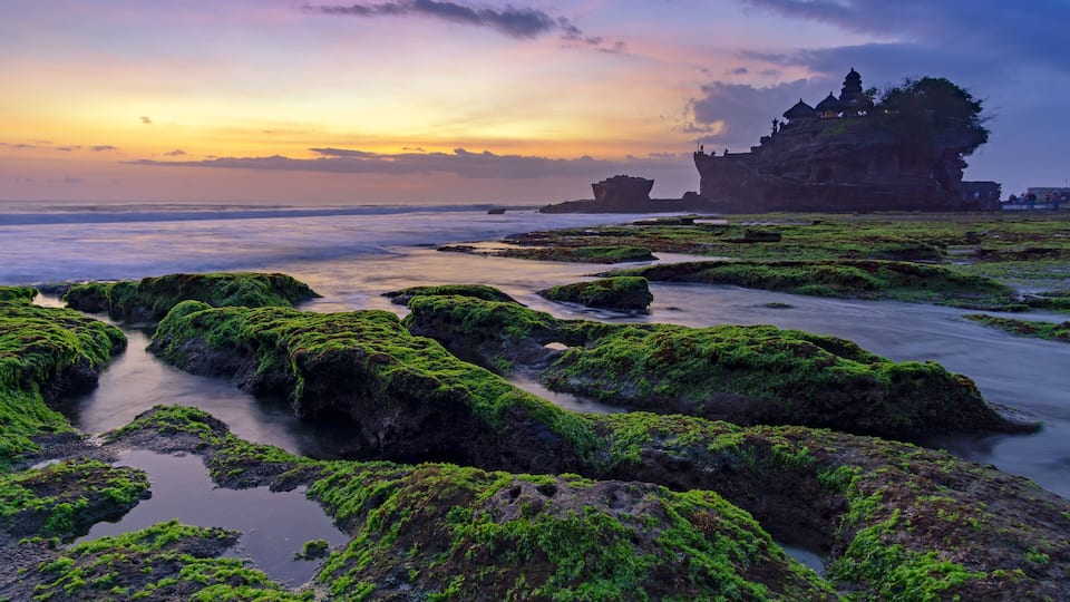 A coastal landscape with green moss-covered rocks, a temple on a cliff, and a sunset in the background.