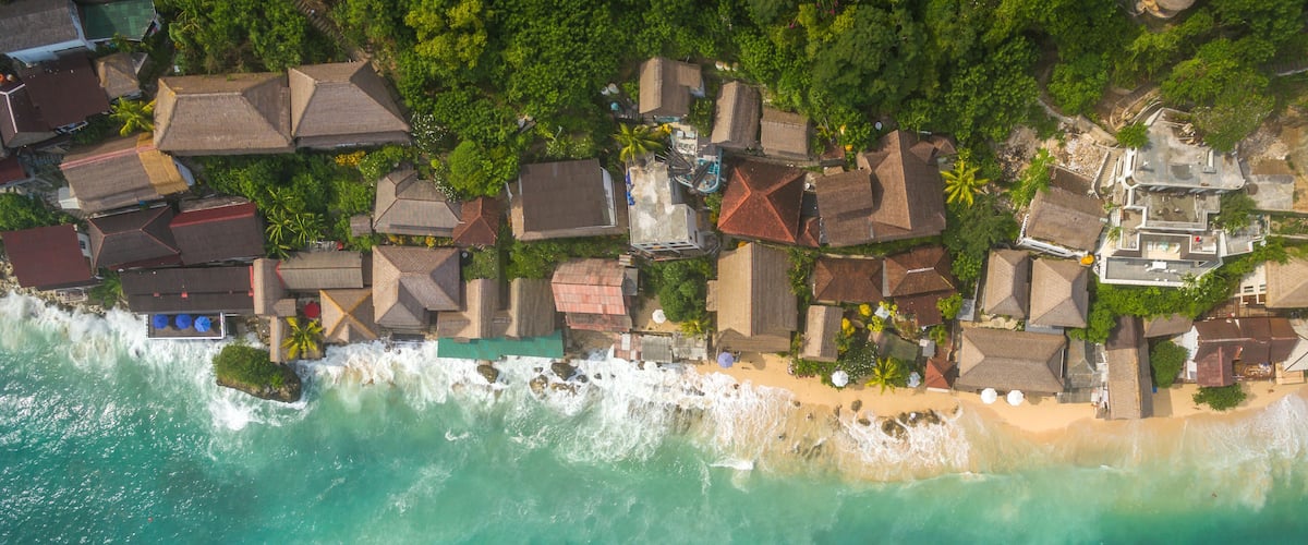 An aerial view of a coastal area with houses, a beach, and waves crashing onto the shore.