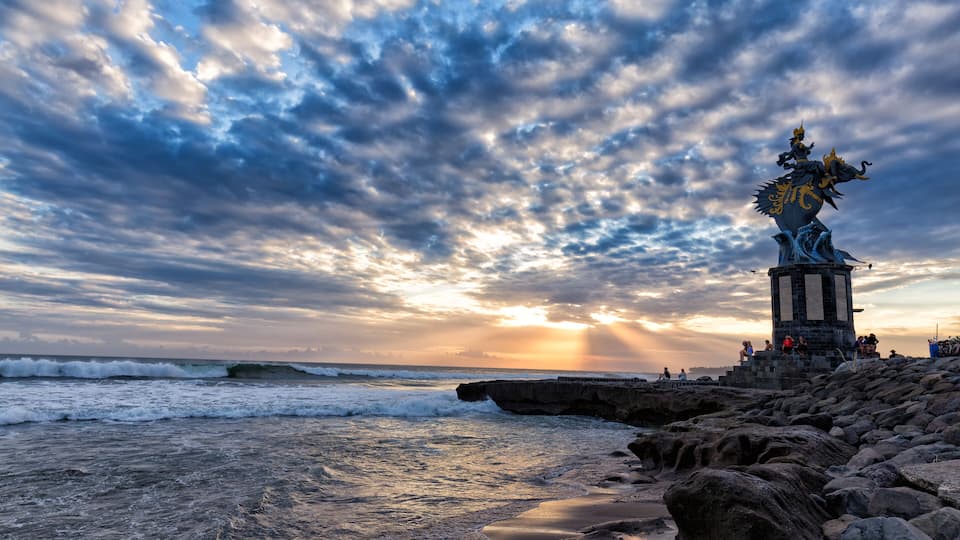 A coastal scene with a statue on a rocky outcrop, waves crashing, and a sunset.