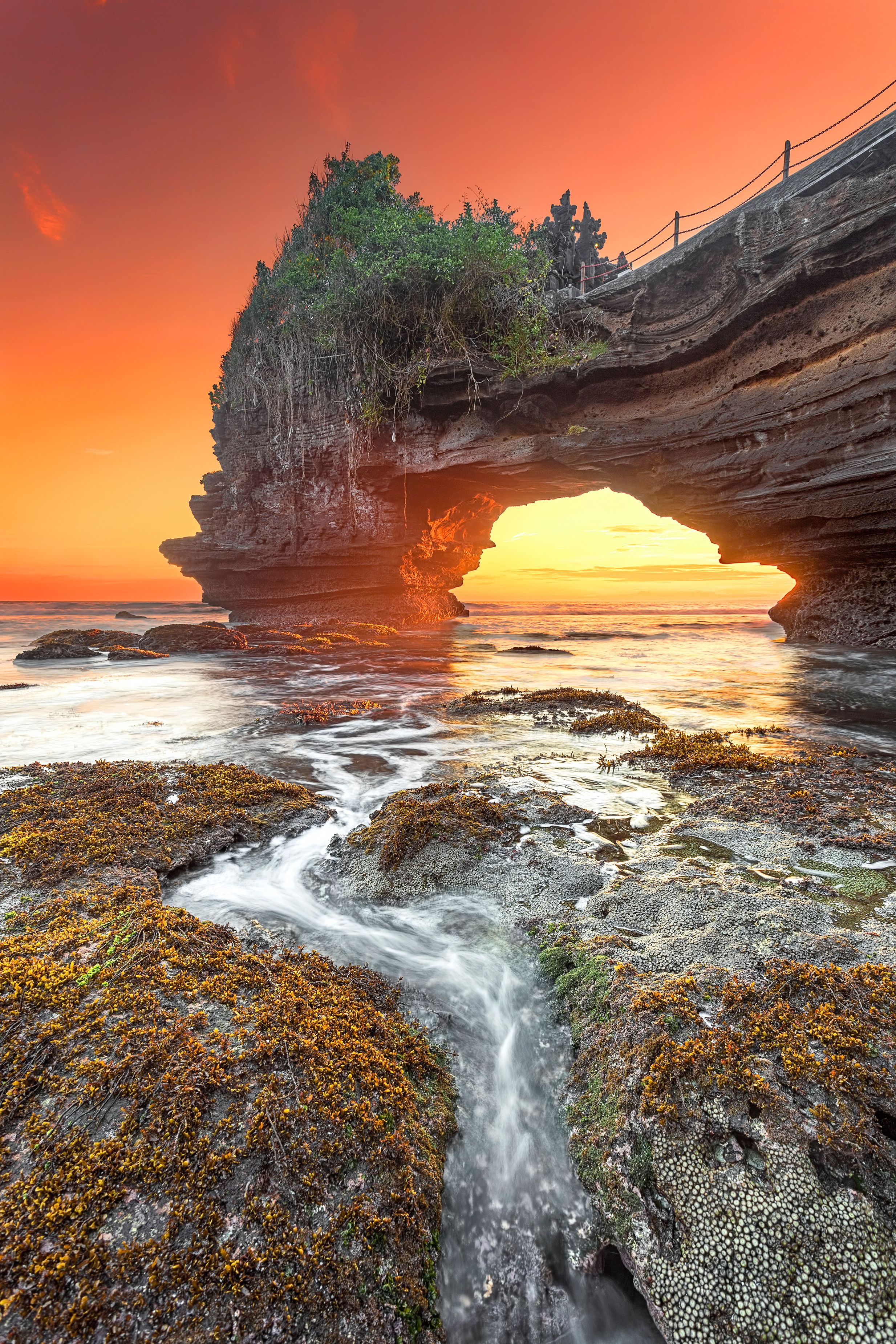 A coastal landscape with a prominent rock formation featuring a natural arch, a flowing stream, and a sunset sky.