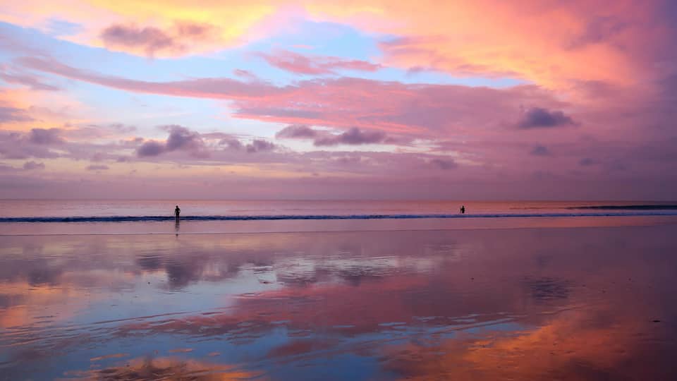A beach with a person standing in the distance, reflecting the colorful sky.