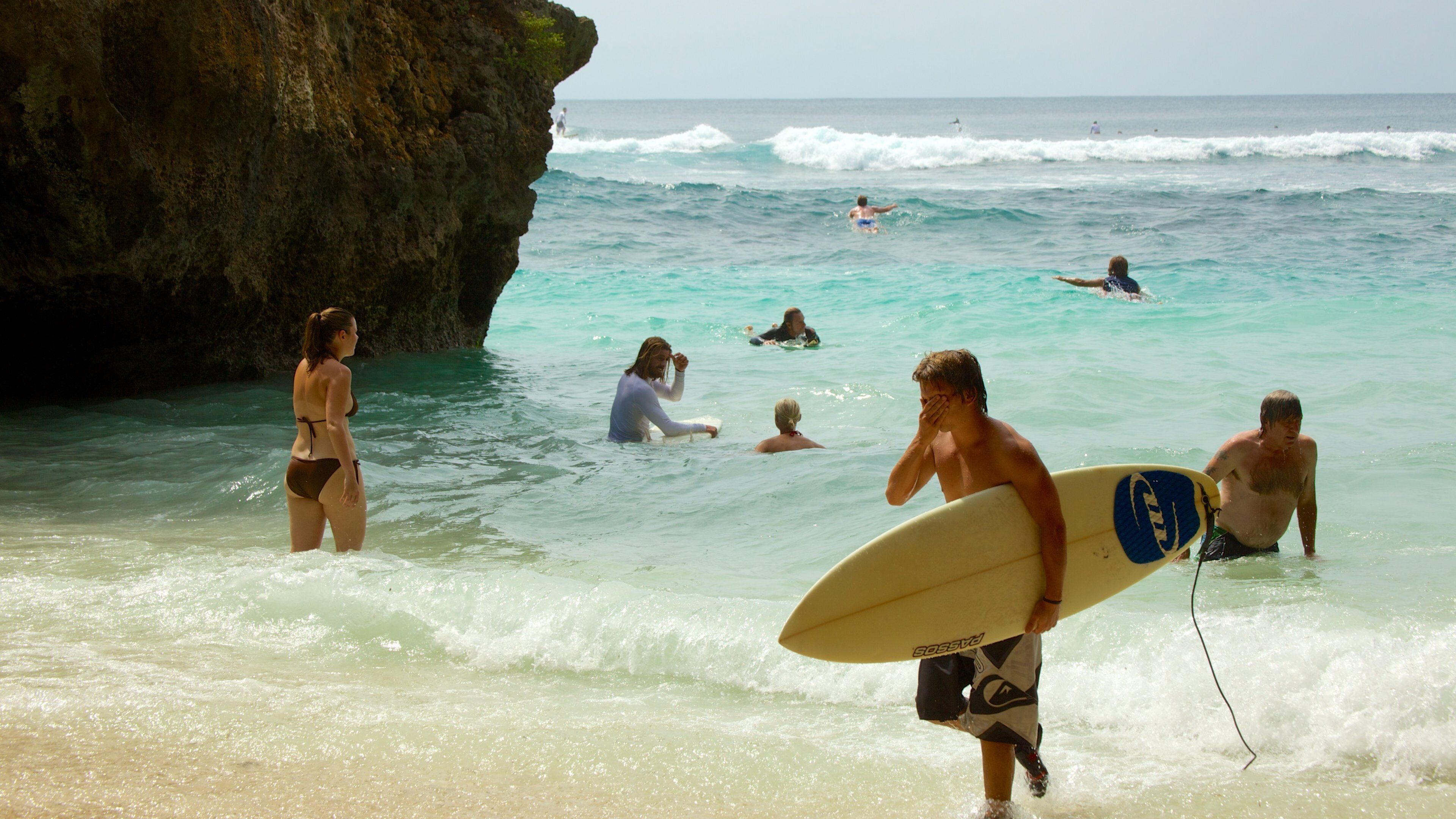 A beach with clear turquoise water, a rocky cliff, and people swimming and surfing.