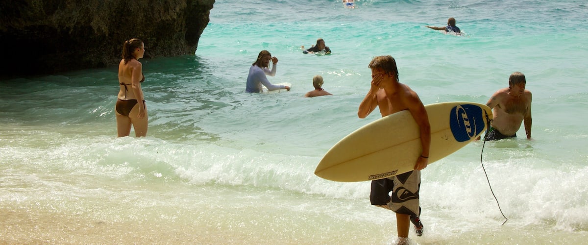 A beach with clear turquoise water, a rocky cliff, and people swimming and surfing.