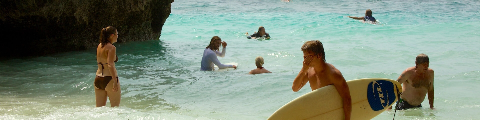 A beach with clear turquoise water, a rocky cliff, and people swimming and surfing.