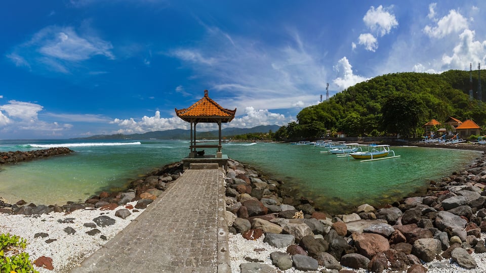 A coastal scene with a gazebo, rocky shoreline, clear turquoise waters, boats, and lush greenery on a hillside.