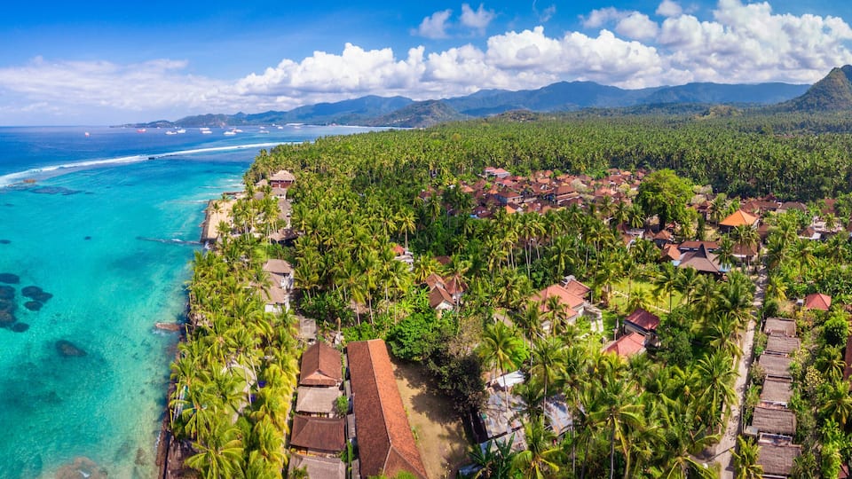 A coastal village with clear turquoise waters, dense palm trees, and a mountain in the background.