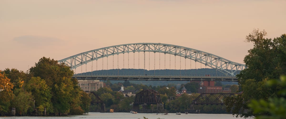 A steel arch bridge over a river with surrounding trees and buildings.