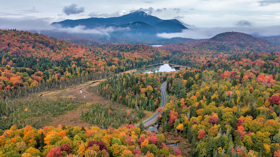 A winding road through a forest with autumn foliage, a lake, and mountains in the background.