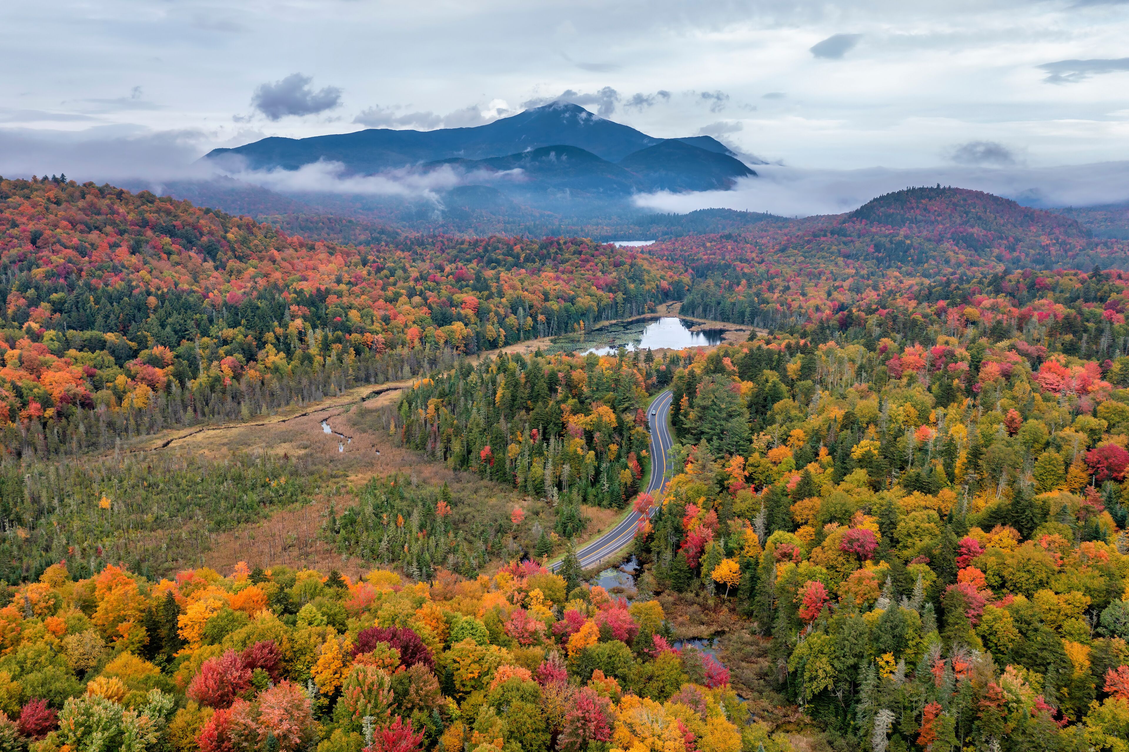 A winding road through a forest with autumn foliage, a lake, and mountains in the background.