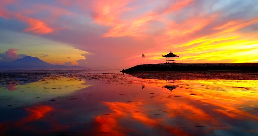 A sunset over a calm sea with a gazebo on a small island and a mountain in the background.
