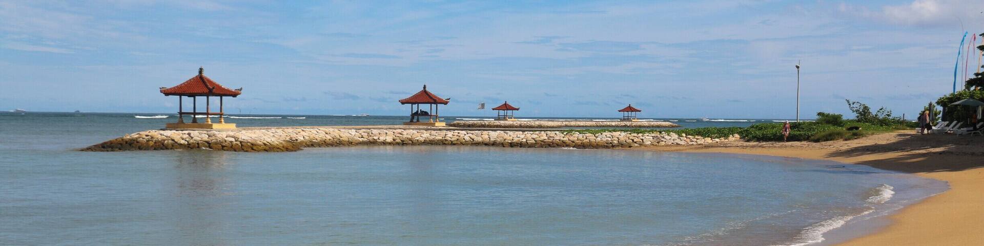 Una playa arenosa con aguas tranquilas, una pequeña isla con construcciones de techo rojo tradicional y un cielo azul despejado.