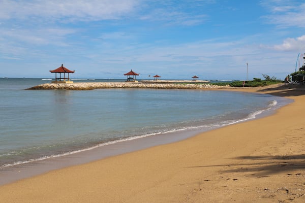 A sandy beach with calm waters, a small island with traditional red-roofed structures, and a clear blue sky.