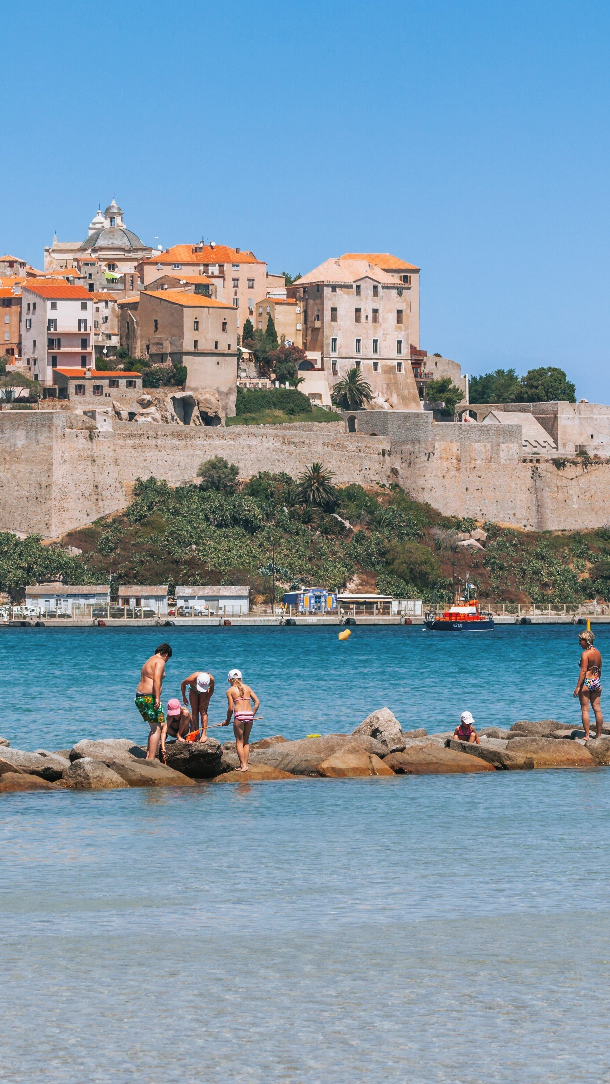 Relaxing summer day at Calvi Beach in Corsica with sunbathers and clear blue waters against historic backdrop