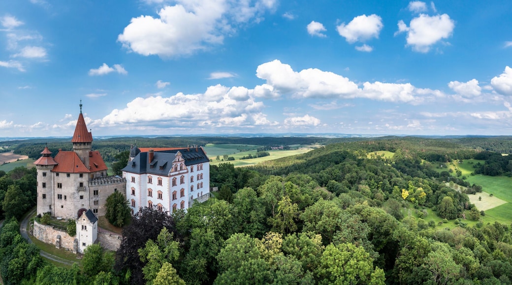 Eine Burg, umgeben von üppigem Grün und sanften Hügeln, unter blauem Himmel.