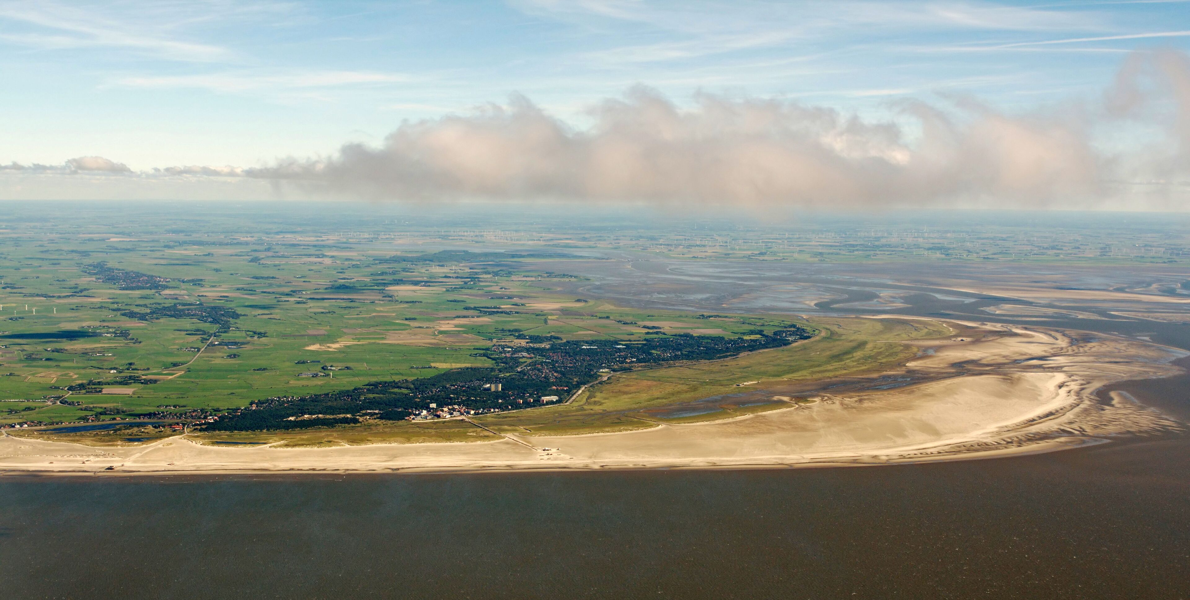 Luftaufnahme einer Küstenlandschaft mit Sandstrand, grünen Feldern und einer Stadt in der Ferne.