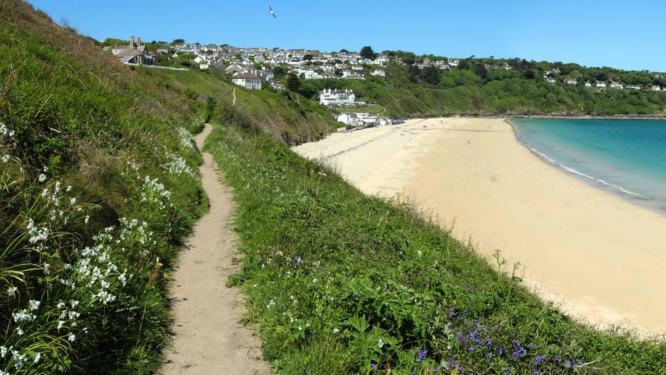 A coastal beach with a sandy shore, a clear blue sea, and a hillside with houses.