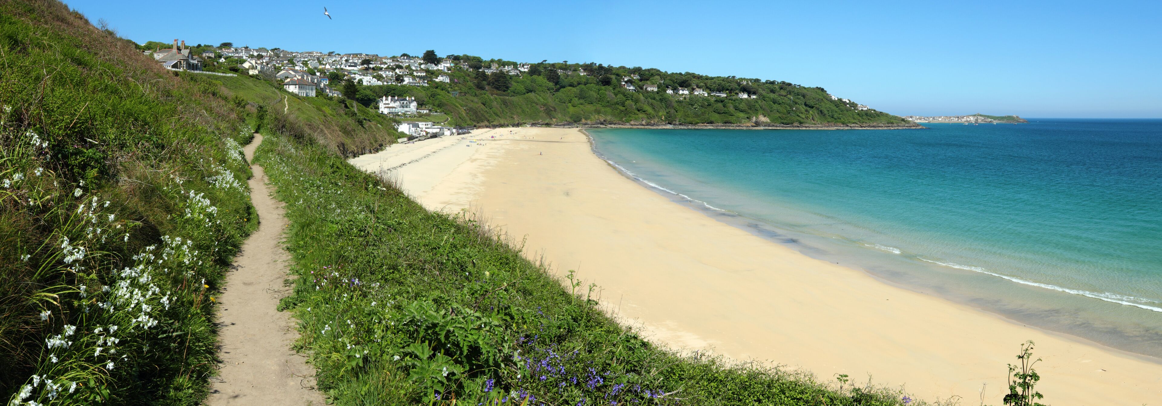 A coastal beach with a sandy shore, a clear blue sea, and a hillside with houses.