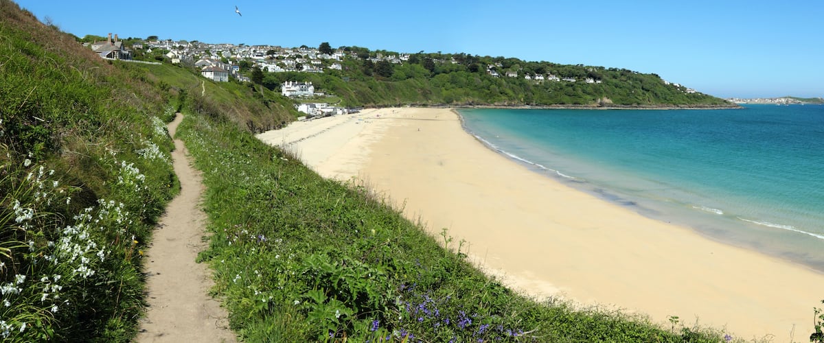 A coastal beach with a sandy shore, a clear blue sea, and a hillside with houses.