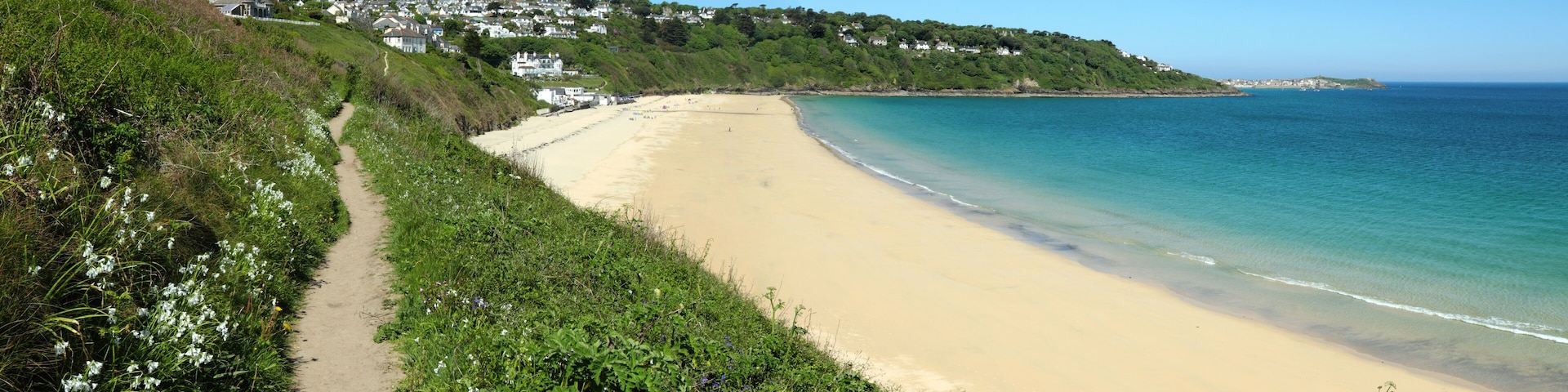 A coastal beach with a sandy shore, a clear blue sea, and a hillside with houses.