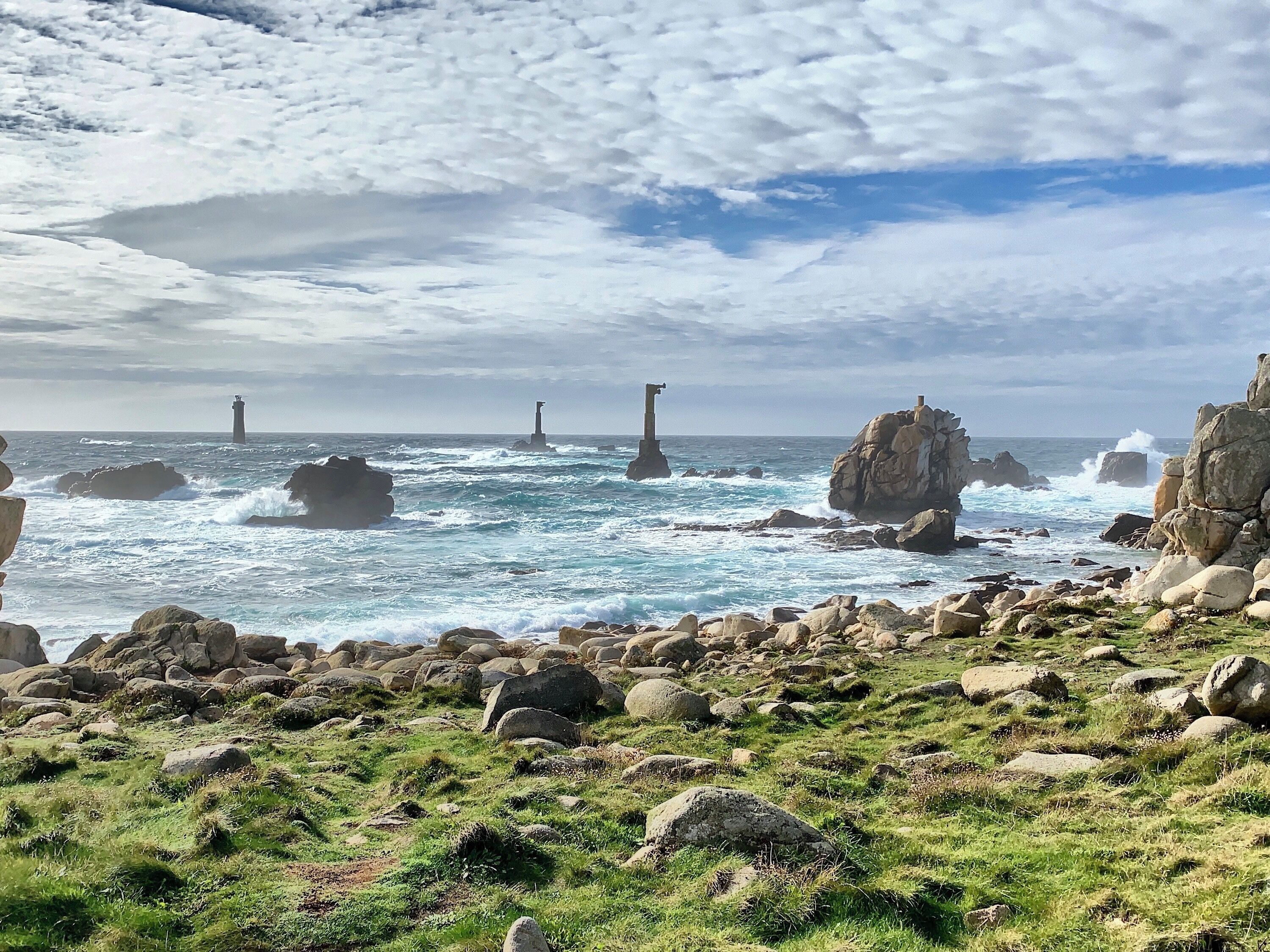 Costa rochosa com ondas quebrando contra grandes pedras e dois faróis à distância.