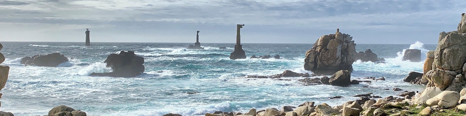 Costa rochosa com ondas quebrando contra grandes pedras e dois faróis à distância.