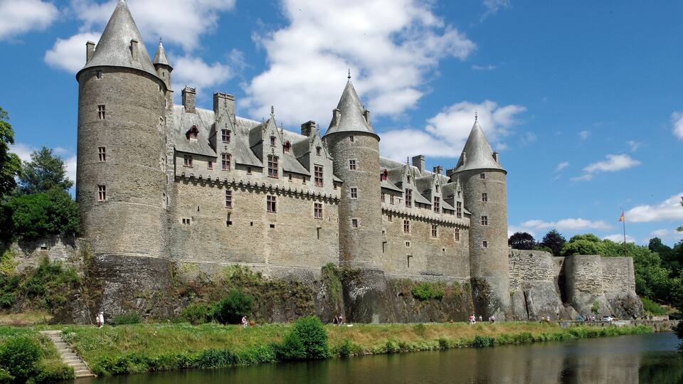 A historic castle with multiple towers, a river in front, and a clear blue sky.
