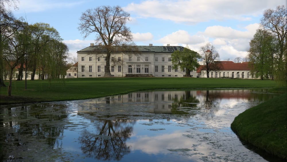 Ein großes, weißes Gebäude mit rotem Dach, umgeben von Bäumen und einem Teich, der das Gebäude spiegelt.