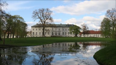 A large white building with a red roof, surrounded by trees and a pond reflecting the structure.