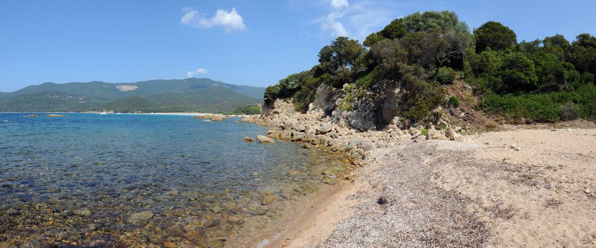 A coastal landscape with clear waters, a sandy beach, rocky outcrops, and dense green trees on a hillside.