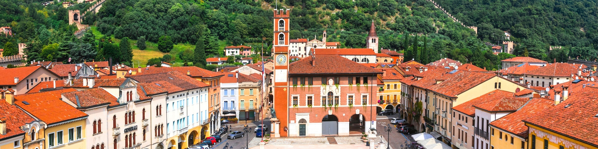 A town square with a large checkered pavement, surrounded by historic buildings with red-tiled roofs. A prominent clock tower is visible in the center, and a hill with a castle-like structure in the background.