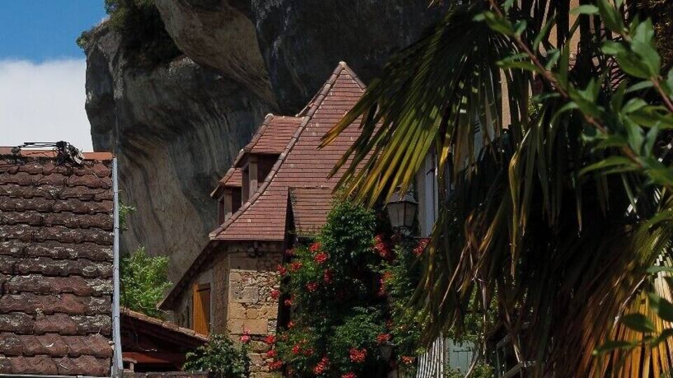 A narrow cobblestone street flanked by stone houses and lush greenery, with a prominent rock formation in the background.